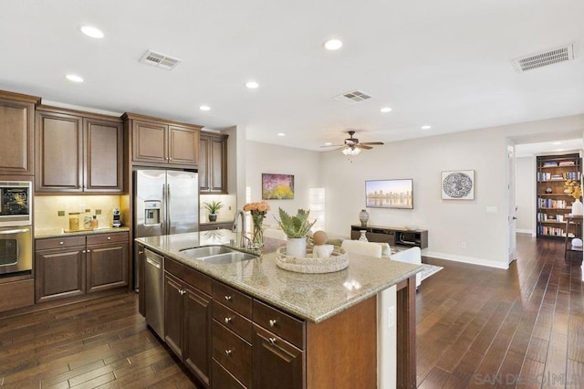 kitchen featuring sink, dark wood-type flooring, stainless steel appliances, light stone countertops, and a center island with sink