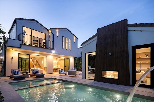 back house at dusk with a balcony, a pool with hot tub, pool water feature, and a patio area