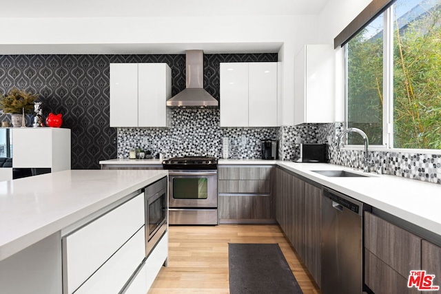 kitchen with sink, white cabinetry, light wood-type flooring, appliances with stainless steel finishes, and wall chimney range hood