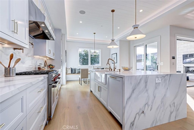 kitchen with an island with sink, white cabinets, high end range, hanging light fixtures, and exhaust hood