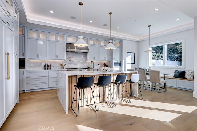 kitchen with decorative light fixtures, decorative backsplash, a tray ceiling, a center island with sink, and light hardwood / wood-style flooring