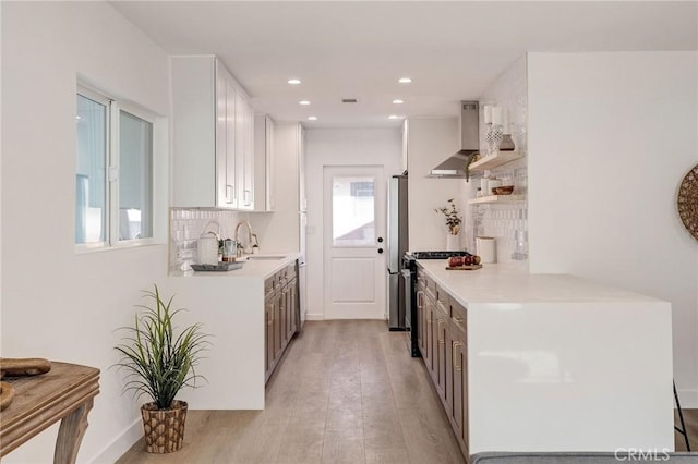 kitchen featuring light wood finished floors, wall chimney exhaust hood, light countertops, open shelves, and backsplash