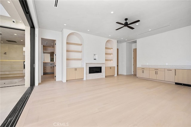 unfurnished living room featuring ceiling fan, a premium fireplace, built in features, and light wood-type flooring