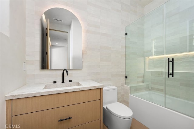 full bathroom with tile walls, bath / shower combo with glass door, vanity, decorative backsplash, and toilet