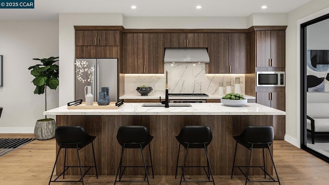 kitchen featuring tasteful backsplash, ventilation hood, a center island with sink, light wood-type flooring, and stainless steel appliances