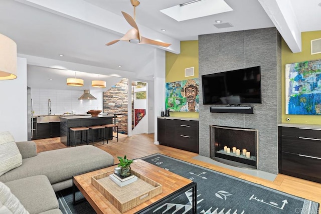 living room with lofted ceiling with skylight, sink, light wood-type flooring, ceiling fan, and a tiled fireplace