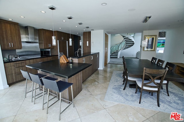 kitchen featuring light tile patterned floors, stovetop, a kitchen island with sink, decorative light fixtures, and wall chimney exhaust hood