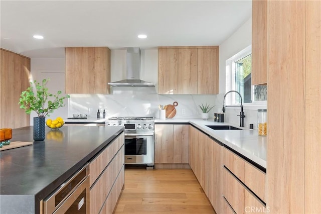 kitchen featuring wall chimney range hood, sink, high end stainless steel range oven, light hardwood / wood-style floors, and light brown cabinetry