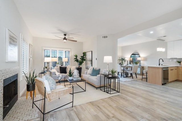 living room with a tiled fireplace, sink, light hardwood / wood-style flooring, and ceiling fan