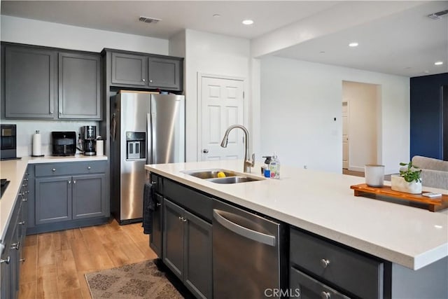 kitchen featuring gray cabinets, an island with sink, sink, stainless steel appliances, and light wood-type flooring