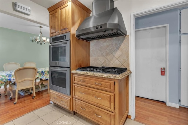 kitchen with light stone counters, decorative backsplash, appliances with stainless steel finishes, ventilation hood, and a chandelier