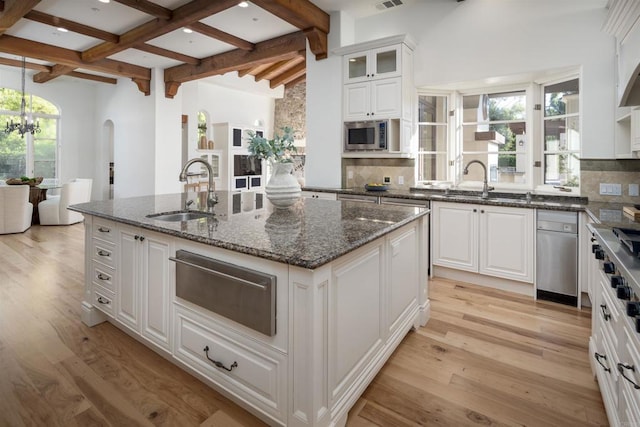 kitchen featuring sink, dark stone countertops, an island with sink, beamed ceiling, and white cabinets