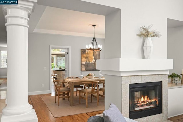 dining space with hardwood / wood-style flooring, a tray ceiling, a tiled fireplace, a chandelier, and ornate columns