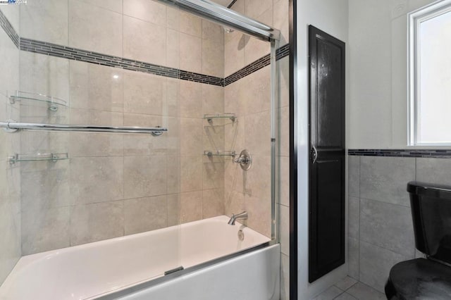 bathroom featuring toilet, a healthy amount of sunlight, bath / shower combo with glass door, and tile walls