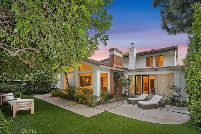 back house at dusk featuring a patio and a lawn