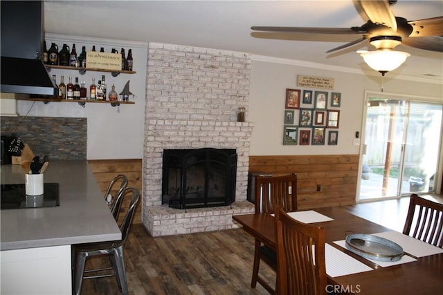 dining room with crown molding, a brick fireplace, dark hardwood / wood-style floors, wooden walls, and ceiling fan