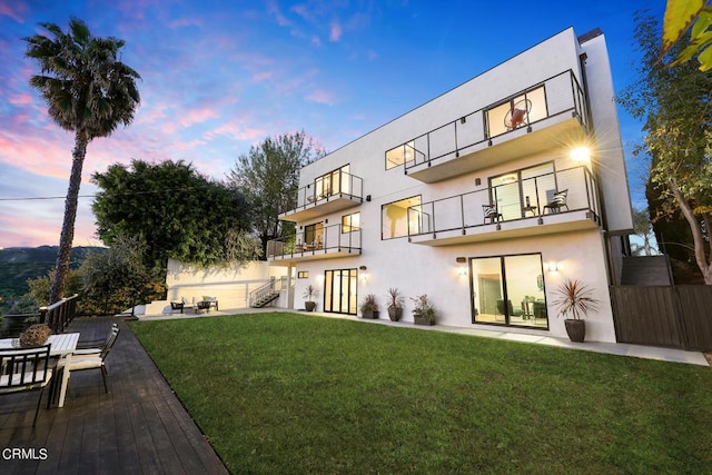 back house at dusk with a balcony, a patio, and a lawn