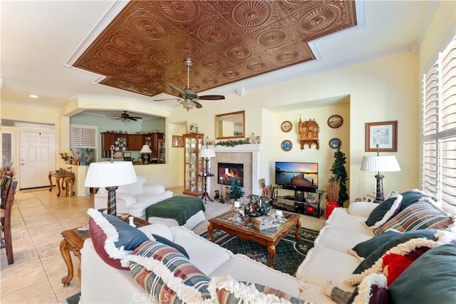 living room with a tiled fireplace, light tile patterned floors, ceiling fan, a raised ceiling, and crown molding