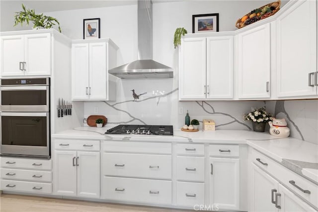 kitchen featuring wall chimney range hood, appliances with stainless steel finishes, white cabinetry, light stone counters, and decorative backsplash