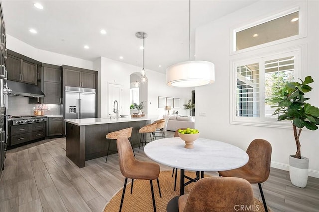 kitchen with a kitchen island with sink, hanging light fixtures, dark brown cabinets, stainless steel appliances, and light hardwood / wood-style floors