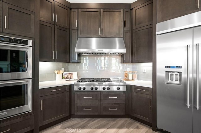 kitchen featuring appliances with stainless steel finishes, backsplash, dark brown cabinetry, extractor fan, and light wood-type flooring