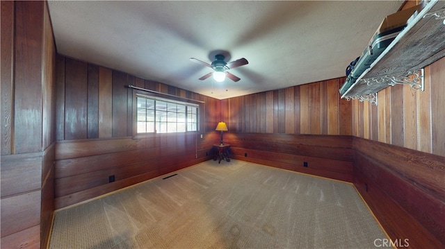 carpeted empty room featuring wooden walls, a ceiling fan, and visible vents