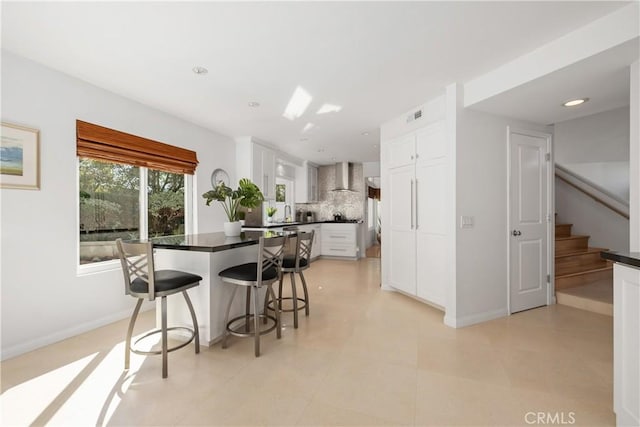 kitchen with white cabinetry, decorative backsplash, wall chimney exhaust hood, and a breakfast bar area