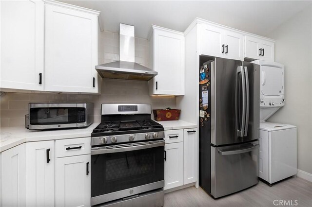 kitchen with stacked washer / dryer, white cabinetry, light countertops, appliances with stainless steel finishes, and wall chimney exhaust hood