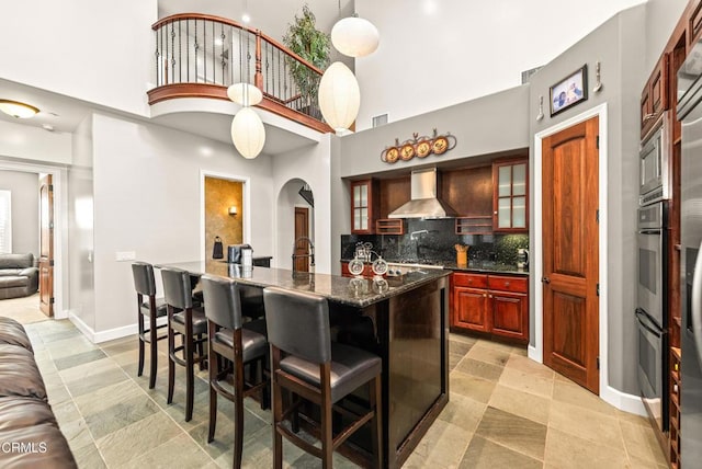 kitchen featuring wall chimney range hood, a breakfast bar, dark stone countertops, backsplash, and a towering ceiling