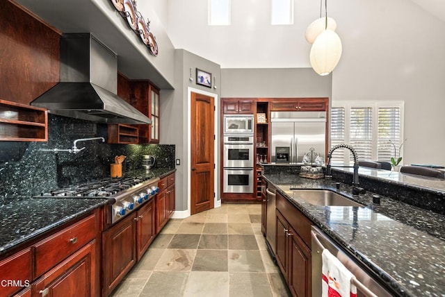 kitchen with pendant lighting, sink, built in appliances, dark stone counters, and wall chimney exhaust hood