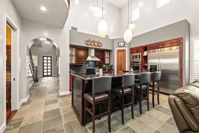 kitchen featuring decorative light fixtures, backsplash, a high ceiling, built in appliances, and wall chimney exhaust hood