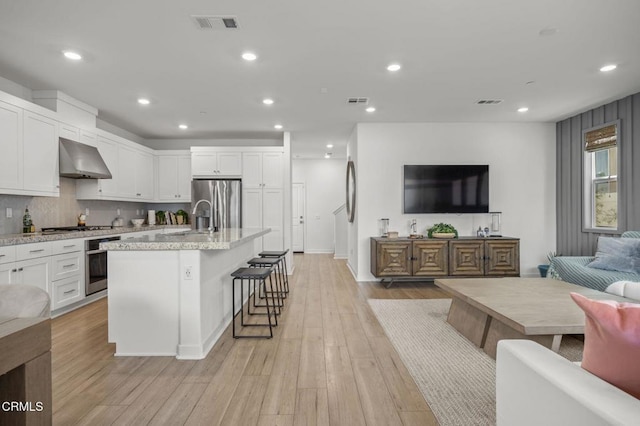 kitchen with wall chimney range hood, a breakfast bar, white cabinetry, stainless steel appliances, and a center island with sink