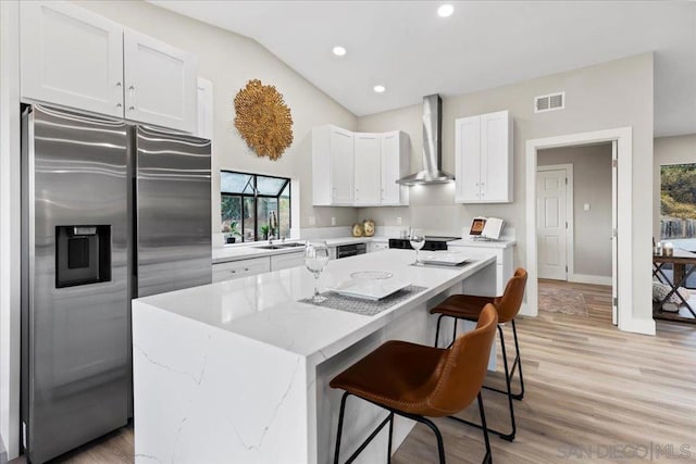 kitchen with white cabinets, stainless steel fridge, a center island with sink, and wall chimney range hood