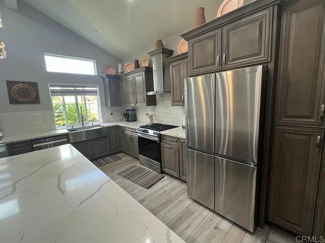 kitchen with wall chimney exhaust hood, light stone counters, light wood-type flooring, stainless steel appliances, and backsplash