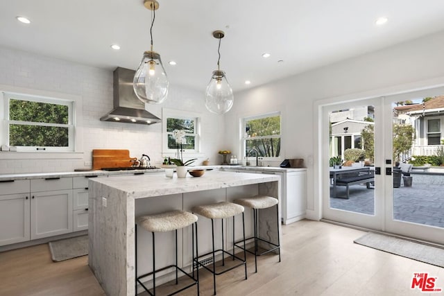 kitchen with pendant lighting, wall chimney range hood, backsplash, a center island, and light stone counters