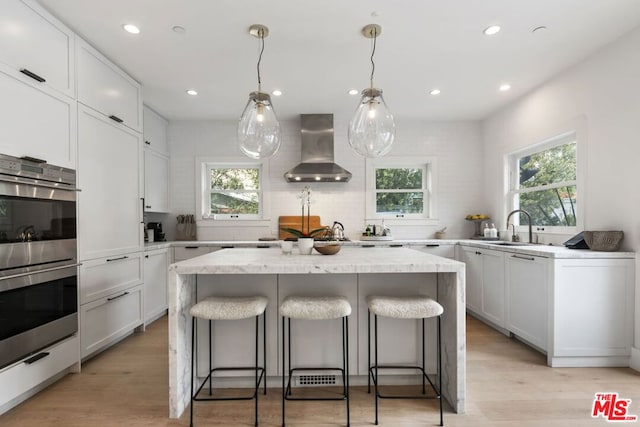 kitchen featuring a kitchen island, double oven, white cabinets, light stone counters, and wall chimney exhaust hood