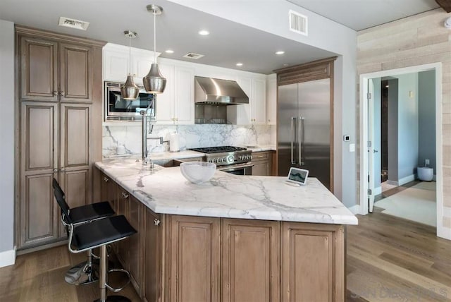 kitchen featuring backsplash, a kitchen bar, a large island with sink, built in appliances, and wall chimney range hood