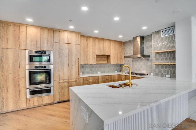 kitchen with sink, wall chimney exhaust hood, kitchen peninsula, light stone countertops, and light hardwood / wood-style flooring