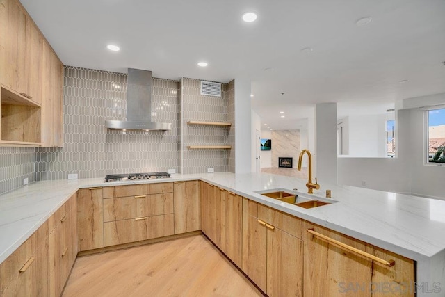 kitchen with light brown cabinetry, sink, stainless steel gas cooktop, kitchen peninsula, and wall chimney range hood