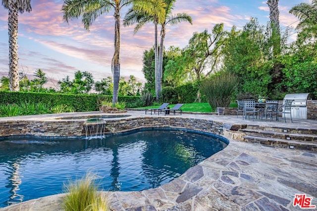 pool at dusk with a patio, area for grilling, exterior kitchen, an in ground hot tub, and pool water feature