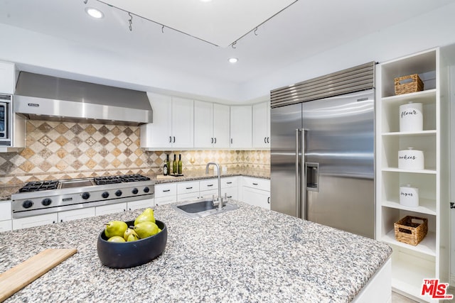 kitchen featuring sink, built in appliances, tasteful backsplash, light stone countertops, and wall chimney exhaust hood