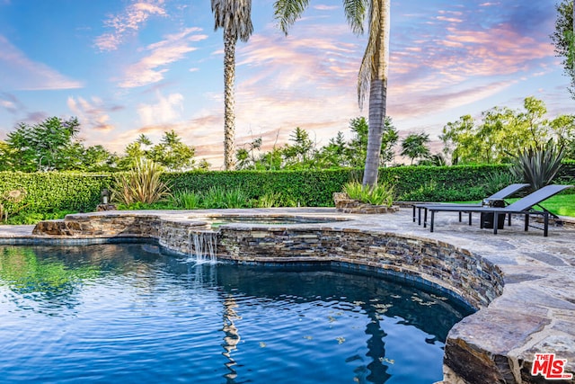 pool at dusk featuring a patio area, pool water feature, and an in ground hot tub