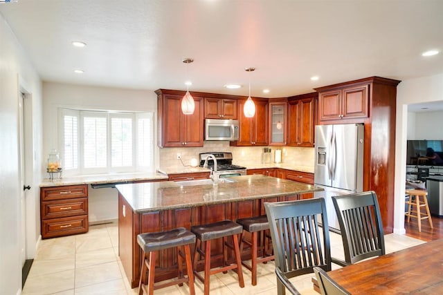 kitchen with light tile patterned floors, appliances with stainless steel finishes, pendant lighting, dark stone counters, and a kitchen island with sink