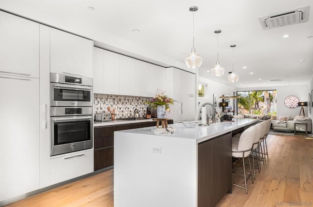 kitchen with white cabinets, a kitchen island with sink, stainless steel appliances, dark brown cabinets, and light wood-type flooring