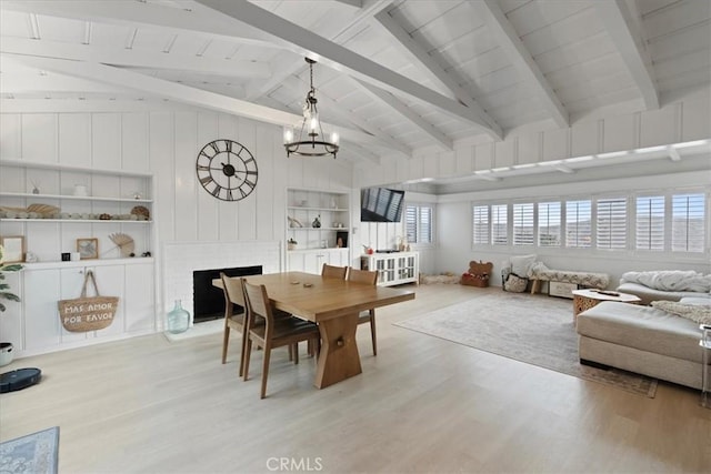 dining space featuring built in shelves, vaulted ceiling with beams, a brick fireplace, light wood-type flooring, and a notable chandelier