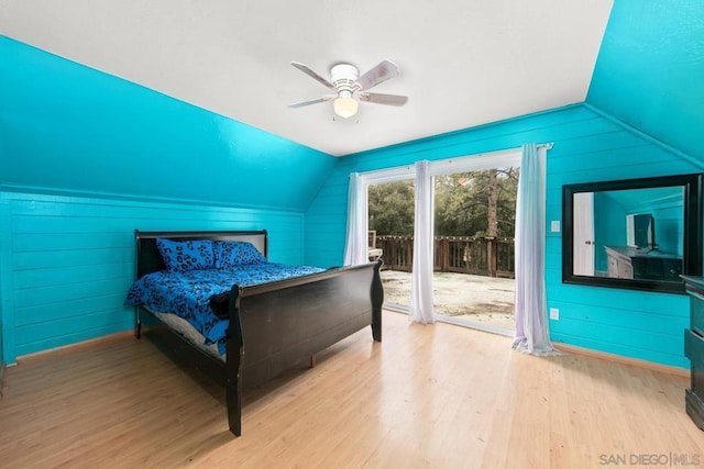 bedroom with vaulted ceiling, wood walls, access to outside, ceiling fan, and light hardwood / wood-style floors