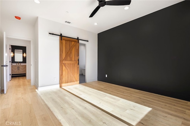 unfurnished bedroom featuring ceiling fan, ensuite bathroom, a barn door, and light hardwood / wood-style flooring