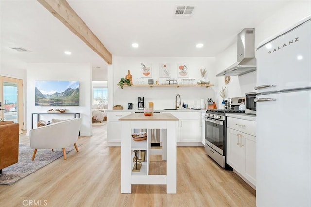 kitchen with appliances with stainless steel finishes, beamed ceiling, white cabinetry, wall chimney exhaust hood, and light hardwood / wood-style flooring