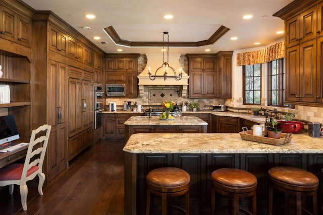 kitchen featuring a center island with sink, stainless steel microwave, dark hardwood / wood-style floors, a tray ceiling, and light stone countertops