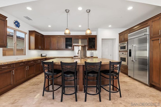 kitchen featuring hanging light fixtures, stainless steel appliances, light stone counters, ventilation hood, and an island with sink